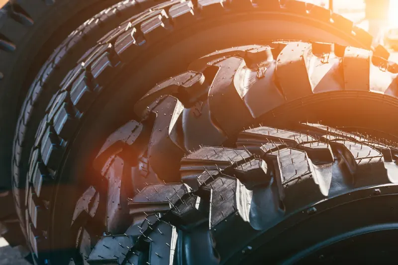 A very close-up shot of the deep, aggressive treads of several large, new tires, with bright sunlight or lens flare creating a warm orange glow across the rubber surfaces.