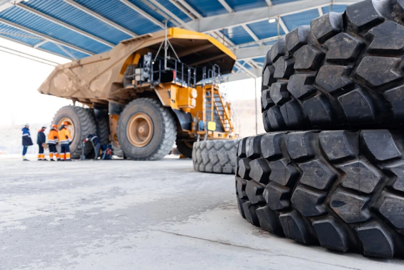 A close-up view of stacked massive off-road tires in the foreground, with a giant yellow haul truck parked under a blue-roofed structure.