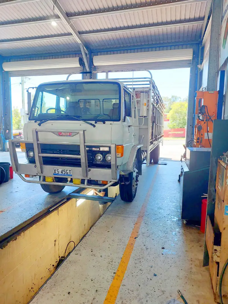 A white Jung truck with a cattle crate or tray body with a large front bull bar over a service pit inside a garage bay.