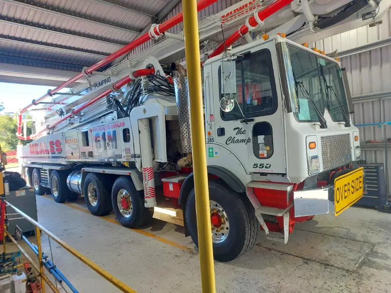 Close-up of a heavy-duty concrete pumping truck with multiple axles, parked indoors under a metal roof, ready for service.